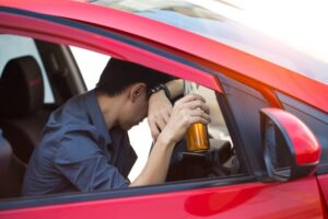 A close-up of a man's hand gripping an alcohol bottle while seated in the driver's seat of a car, illustrating the dangerous act of drinking while driving.
